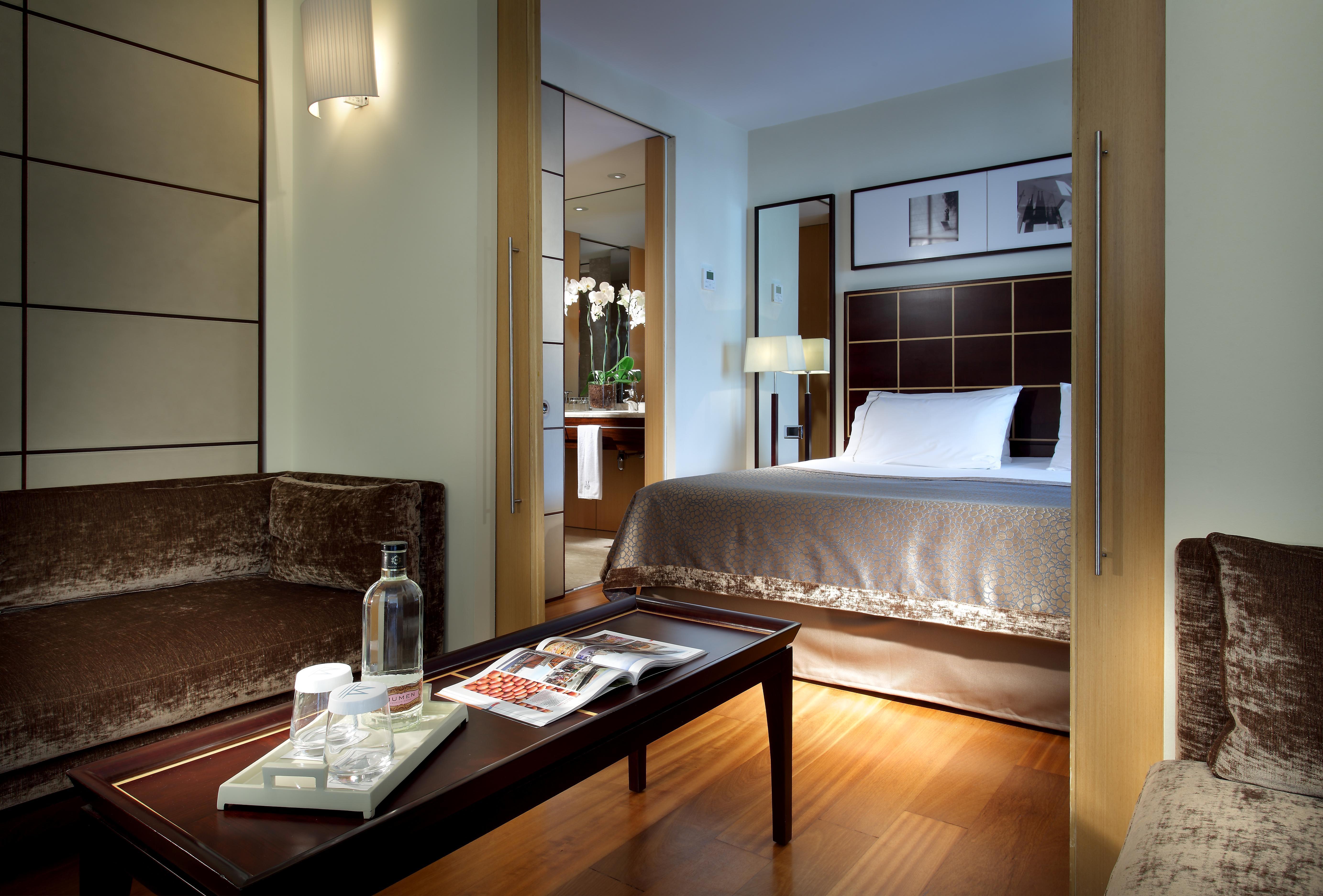 Eurostars Grand Marina Hotel Gl Barcelona Ngoại thất bức ảnh The image shows a well-decorated hotel room featuring a comfortable bed with a plush headboard, complemented by a subtle color palette. There is a cozy seating area with a brown couch and a coffee table. On the table, there are items like a water bot