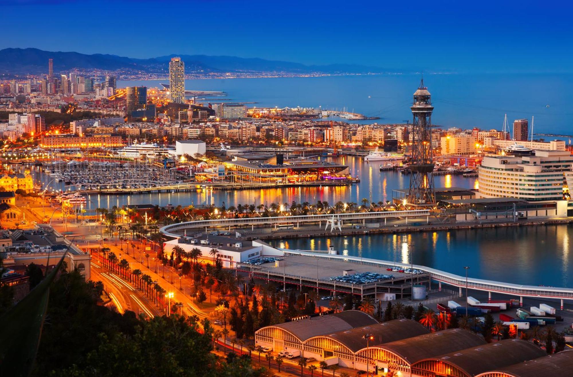 Eurostars Grand Marina Hotel Gl Barcelona Ngoại thất bức ảnh The photo depicts a scenic view of a coastal city at dusk. In the foreground, there are illuminated buildings and a marina filled with boats. The waterfront features a curved walkway and a dock area. In the background, you can see a skyline with tall