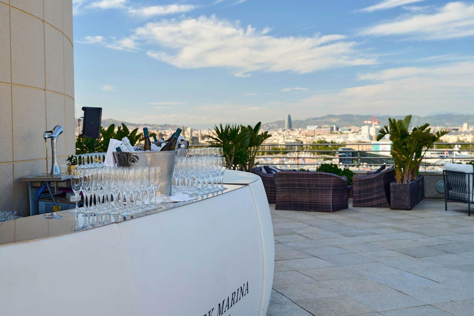 Eurostars Grand Marina Hotel Gl Barcelona Ngoại thất bức ảnh The photo shows a stylish rooftop bar or terrace area with a modern, curved white bar counter. In front of the bar, there is a row of sparkling glasses lined up for drinks. The background features a beautiful skyline and distant mountains, with a cle