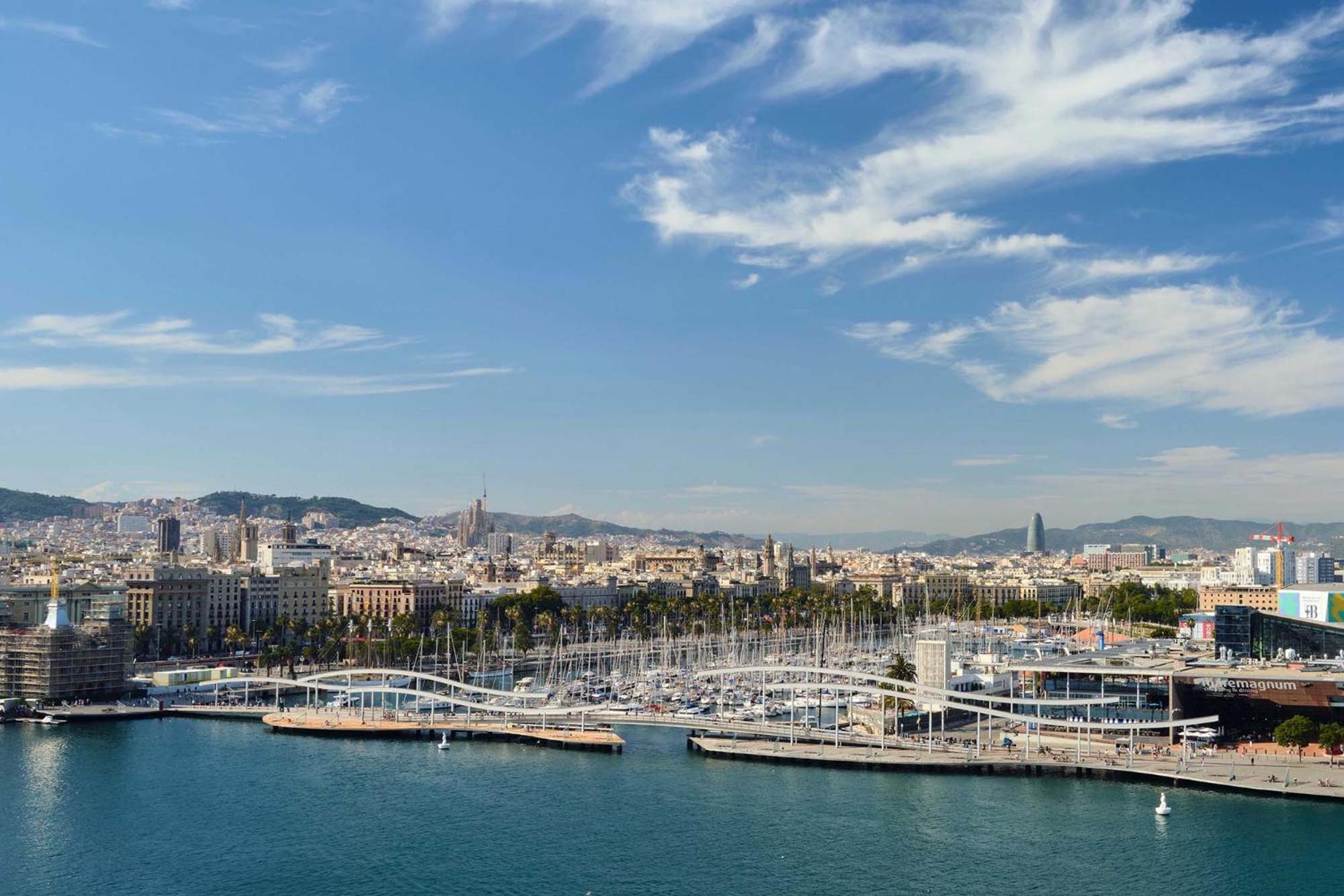 Eurostars Grand Marina Hotel Gl Barcelona Ngoại thất bức ảnh The photo shows a scenic view of a coastal city, likely Barcelona, with a harbor area prominent in the foreground. There are several boats moored along the docks, and a distinctive bridge or walkway structure can be seen connecting different parts of