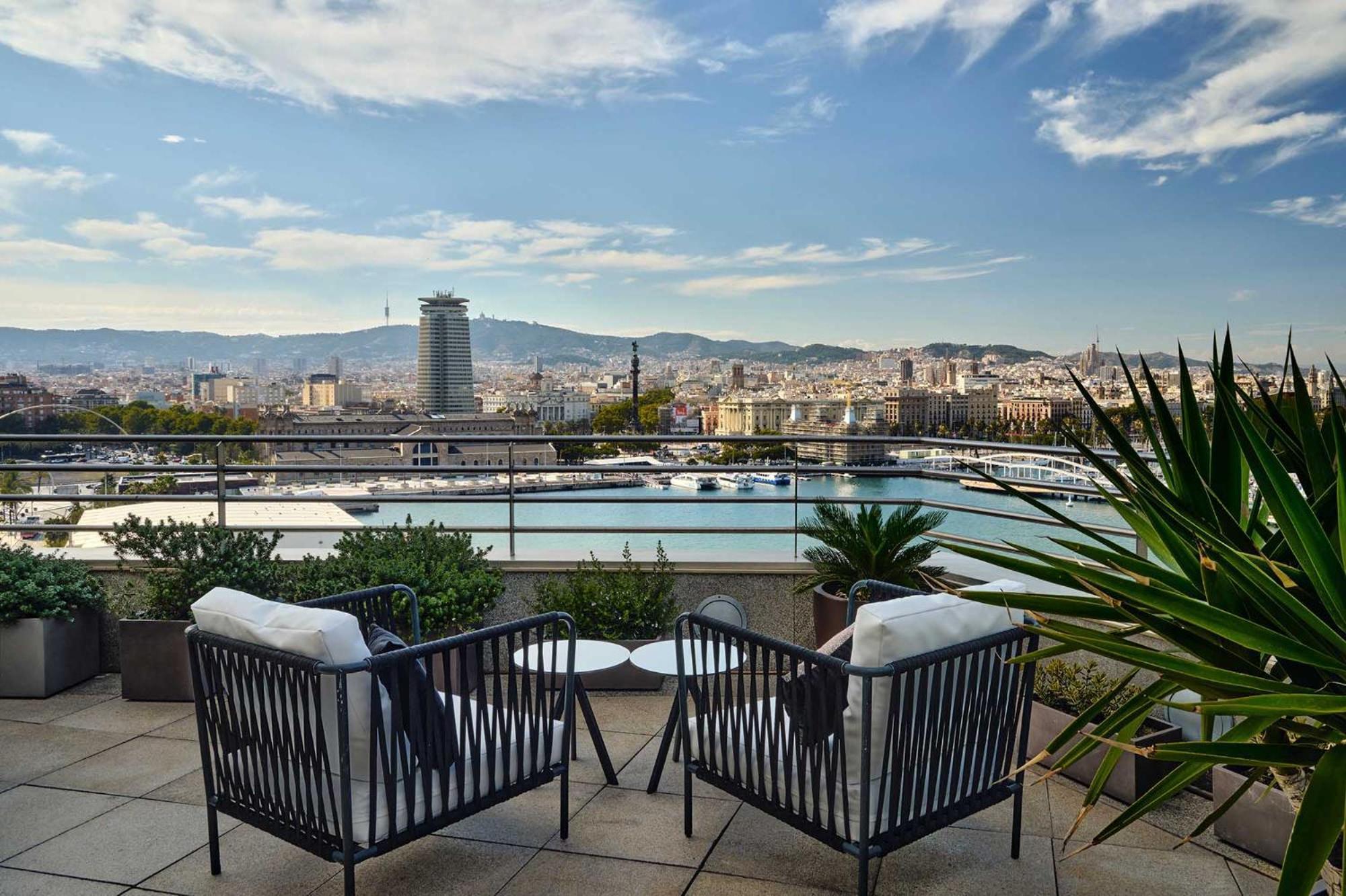 Eurostars Grand Marina Hotel Gl Barcelona Ngoại thất bức ảnh The photo shows a scenic outdoor terrace area featuring two stylish black chairs with white cushions. In the background, there’s a beautiful view of a cityscape with a harbor and mountains in the distance. The sky is partly cloudy, and there are some
