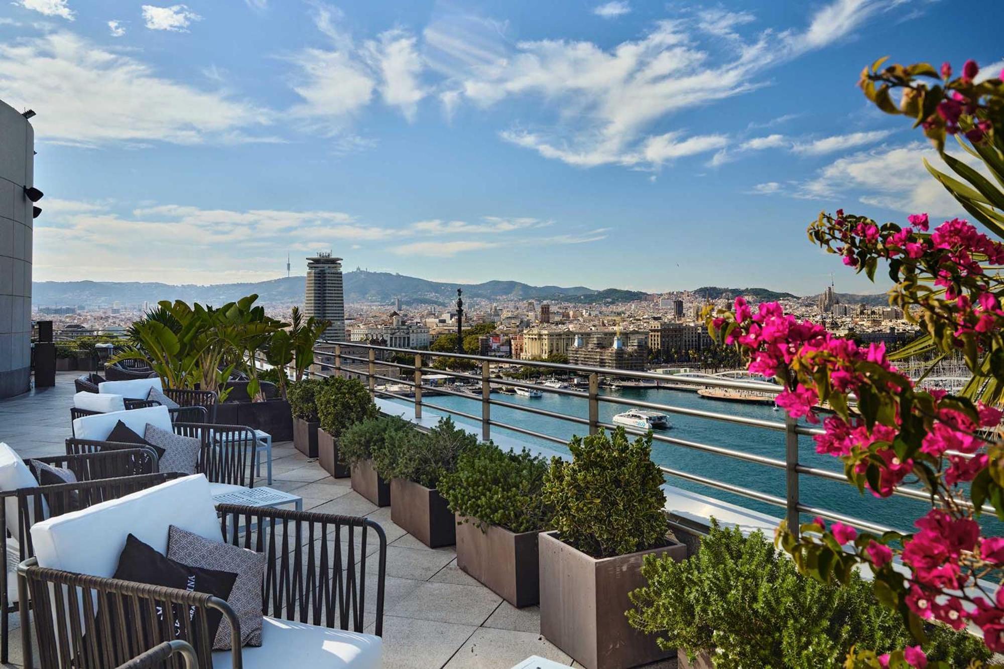 Eurostars Grand Marina Hotel Gl Barcelona Ngoại thất bức ảnh The photo depicts a rooftop terrace with a stunning view of a city, likely Barcelona. There are comfortable seating arrangements, including dark outdoor chairs and cushions, surrounded by decorative plants in large pots. Colorful flowers, possibly bo