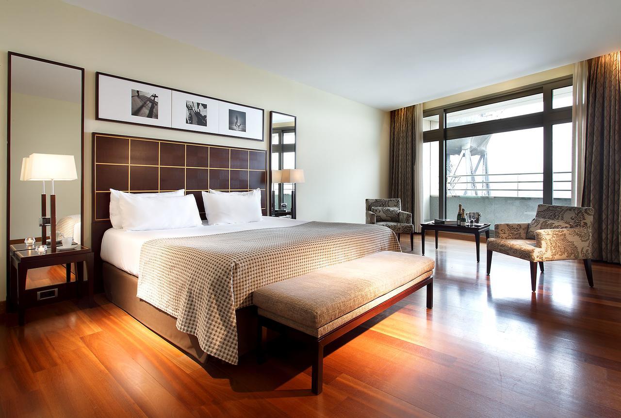 Eurostars Grand Marina Hotel Gl Barcelona Ngoại thất bức ảnh The photo shows a modern hotel room featuring a large bed with a decorative headboard. There are two bedside lamps on either side of the bed, and behind the bed, there are framed pictures hanging on the wall. A bench sits at the foot of the bed, and 