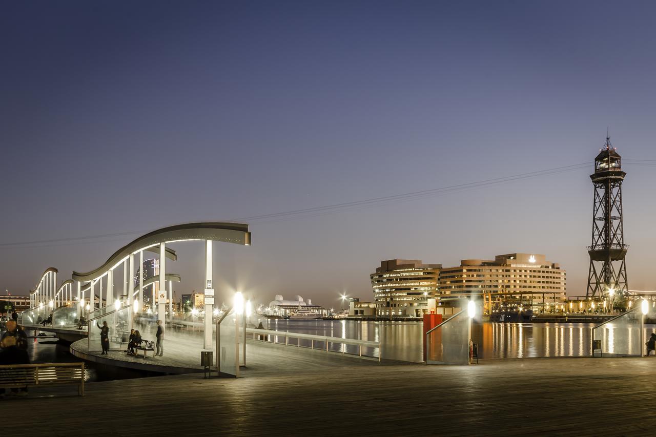 Eurostars Grand Marina Hotel Gl Barcelona Ngoại thất bức ảnh The photo depicts a scenic waterfront view during twilight. On the left, there is a modern, curved pedestrian walkway that appears to be well-lit, inviting pedestrians. To the right, a tall structure resembling a lighthouse or observation tower stand